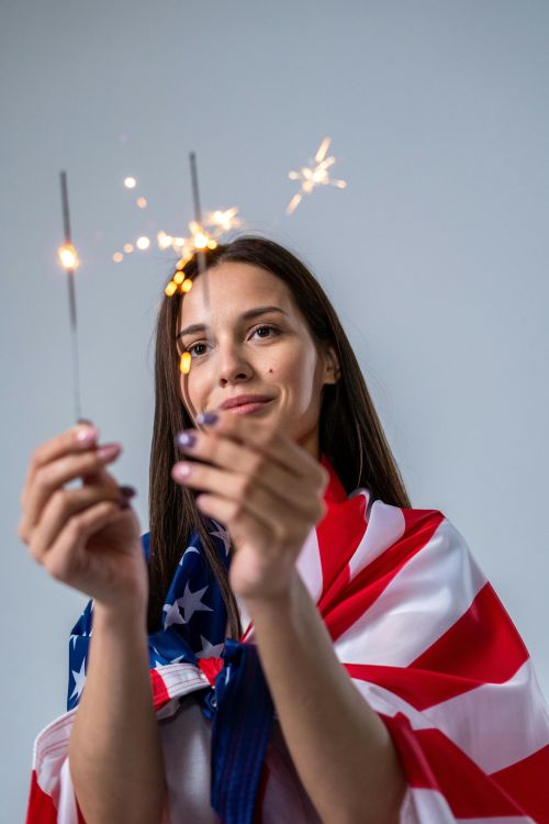 A young woman wrapped in an American flag holding sparklers, symbolizing Independence Day celebrations.