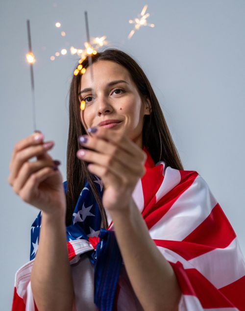 A young woman wrapped in an American flag holding sparklers, symbolizing Independence Day celebrations.